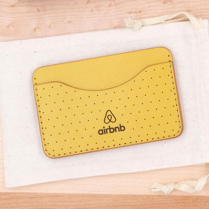 Yellow leather Slim Wallet front view with decorative perforation, Airbnb logo, and canvas bag.